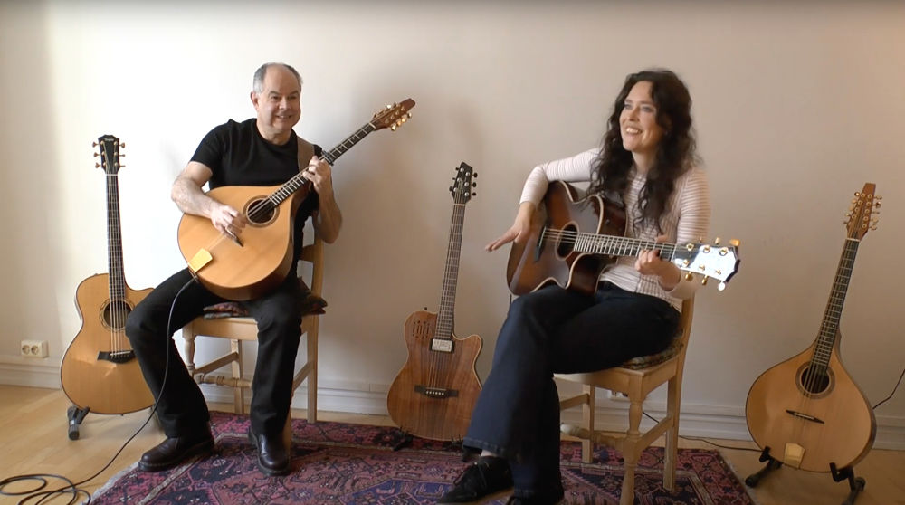 Andy and Viil smiling whilst sitting on chairs and playing guitar and bouzouki