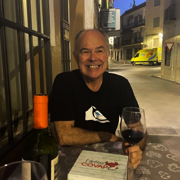 Andy sitting in a pavement restaurant at dusk with a bottle of wine in front of him. 